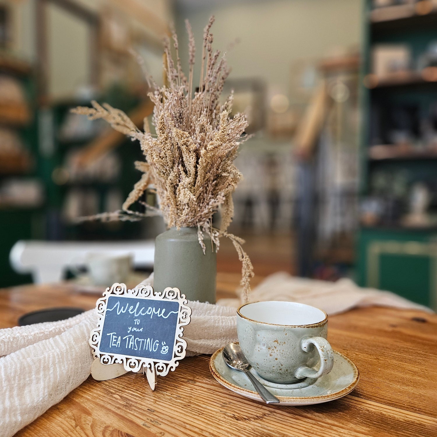 A styled tabletop scene in a shop or cafe with a small ceramic teacup and saucer, a vase of dried grasses, a small welcome sign reading 'Welcome to Tea Tasting', and a folded cream cloth on a wooden table. The background is softly blurred with shelves and interior decor visible.